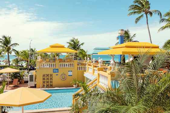 Et tropisk miljø med et basseng og gule parasoller, omgitt av palmer med havet i bakgrunnen. (AI-generert tekst)