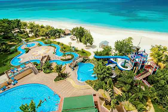 Bildet viser en strand foran et feriested med bassenger, vannsklier og omkringliggende vegetasjon. (AI-generert tekst)