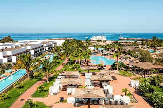 Ferieanlegg med hvite bygninger, et svømmebasseng og grøntområde rundt, samt hav i bakgrunnen. (AI-generert tekst)