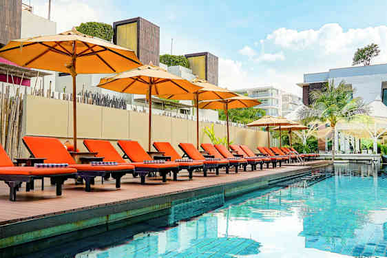 Et utendørsbasseng med oransje solsenger og parasoller ved kanten, omgitt av moderne arkitektur. (AI-generert tekst)