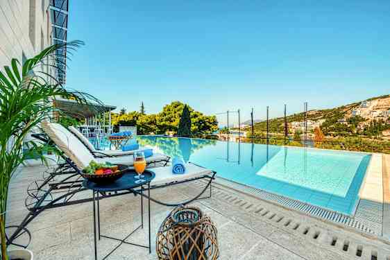 En utsikt mot et infinitybasseng med solsenger, bord med frukt og drikke ved siden av, samt grønne omgivelser og himmel i bakgrunnen. (AI-generert tekst)