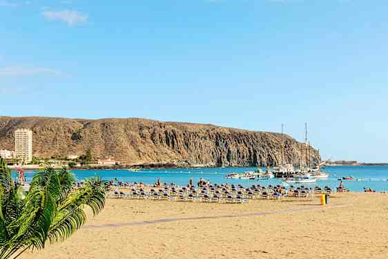 Strand med solsenger, havet og båter ved en klippe. (AI-generert tekst)