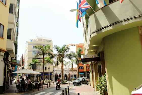 En gate med bygninger, palmer og et hotellskilt. Folk sitter ved en kafé, og et motorkjøretøy passerer. (AI-generert tekst)