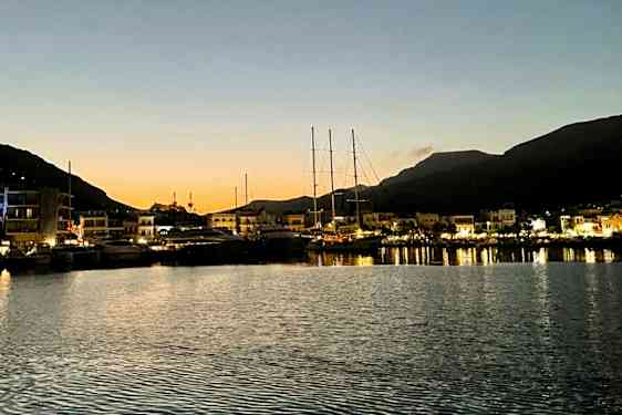 En havn i skumringen med båter fortøyd ved en kai og opplyste bygninger i bakgrunnen. Fjell kan sees i bakgrunnen. (AI-generert tekst)