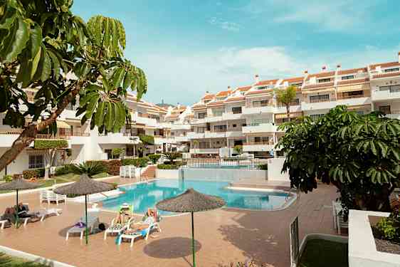 Et utendørsområde med et basseng, solsenger og et fleretasjehotell. Flere parasoller og grønne planter omgir området. (AI-generert tekst)