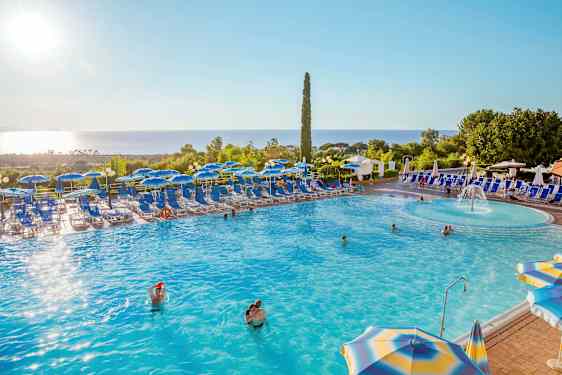 Et svømmebasseng med solsenger og parasoller rundt, omgitt av grøntområder og med havutsikt. (AI-generert tekst)