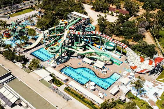 Et vannland med flere store vannsklier, bassenger og solsenger, omgitt av grønne trær og bygninger. (AI-generert tekst)