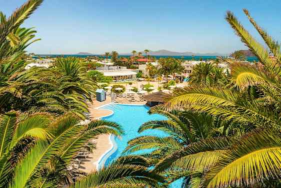 En utsikt over et svømmebasseng omgitt av palmer med havet i det fjerne. (AI-generert tekst)