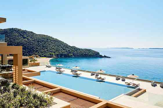 Utsikt over et basseng med solsenger og parasoller, en kystlinje og hav i bakgrunnen. (AI-generert tekst)