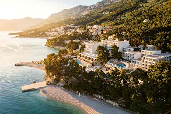 Luftfoto av hotell ved kysten og strand, omgitt av trær og med fjell i bakgrunnen. (AI-generert tekst)