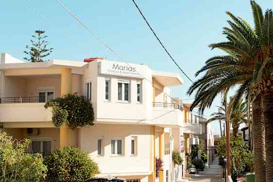 Eksteriør av en bygning med skiltet "Marias Studios & Apartments" og omkringliggende tropiske planter. (AI-generert tekst)