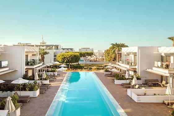 Hotellområde med et langt basseng omgitt av solsenger og parasoller, med hvite bygninger i bakgrunnen. (AI-generert tekst)