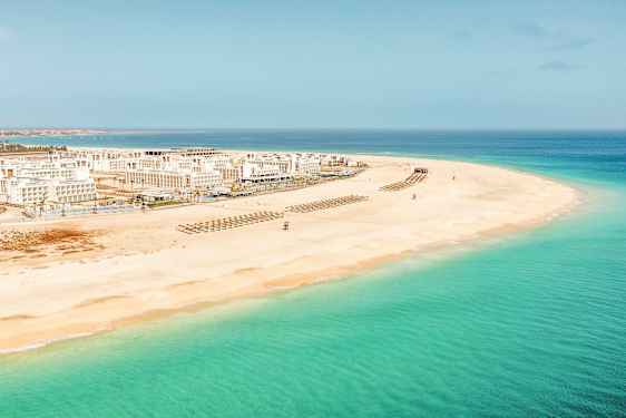 Luftfoto av en hvit sandstrand med klart turkist vann og hvite bygninger i bakgrunnen. (AI-generert tekst)