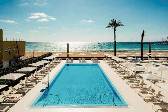 Et svømmebasseng omgitt av solsenger og parasoller nær en sandstrand og hav. (AI-generert tekst)