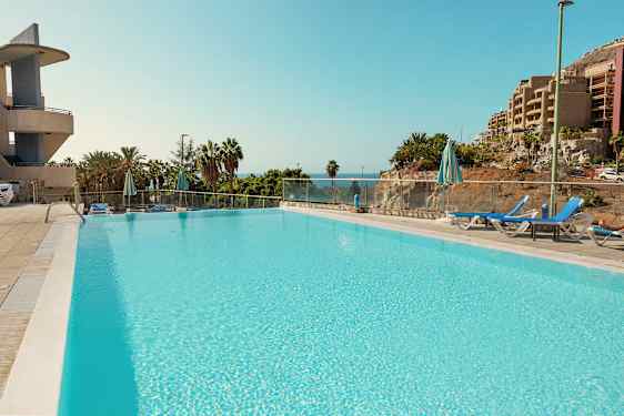 Et stort basseng omgitt av solsenger og parasoller, med utsikt mot havet og frodig vegetasjon. (AI-generert tekst)