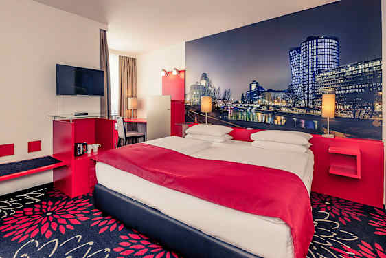 Et hotellrom med en dobbeltseng, en TV på veggen og et skrivebord ved siden av vinduet. Veggene er dekorert med et storbilde av et bylandskap. (AI-generert tekst)