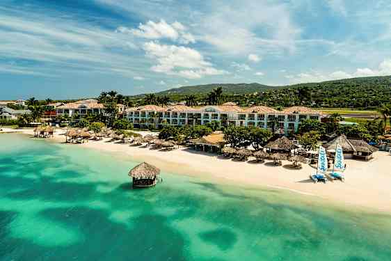 Resort ved strand med grøntblått vann, palmer og bygninger. (AI-generert tekst)