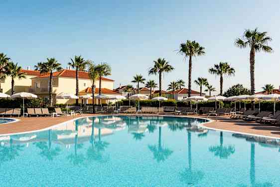 Utendørsbasseng med solsenger og parasoller, omgitt av palmer og bygninger med terrakottatak. (AI-generert tekst)