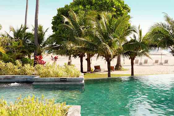 Bassengområde med utsikt mot en sandstrand og omkringliggende palmer. (AI-generert tekst)