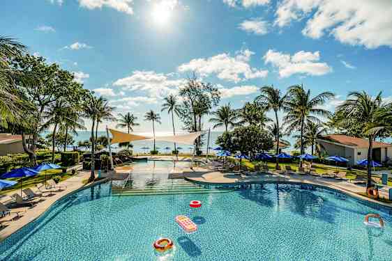 Et svømmebasseng omgitt av solsenger og parasoller, med utsikt over havet og palmer under en klar himmel. (AI-generert tekst)