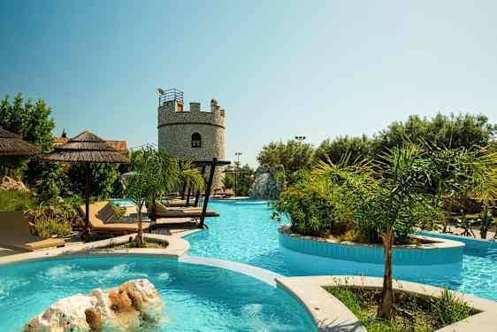 Et utendørs svømmebasseng med solsenger og parasoller, og en steinbygning i bakgrunnen. (AI-generert tekst)