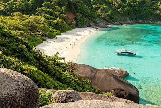 En sandstrand med mennesker, omgitt av frodige trær og klart turkis vann. En båt ligger nær stranden. (AI-generert tekst)