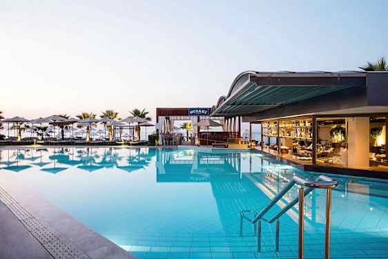 Et stort basseng ved en hotellrestaurant med utsikt over havet. Solstoler og parasoller omgir området. (AI-generert tekst)