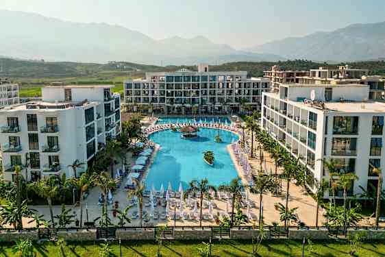 Ett moderne hotellanlegg med et stort basseng og solsenger rundt, plassert i en frodig dal med fjell i bakgrunnen. (AI-generert tekst)