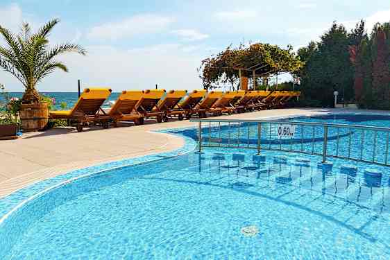 Et svømmebasseng med grunt vann foran en rekke gule solsenger på en terrasse, med havet i bakgrunnen. (AI-generert tekst)