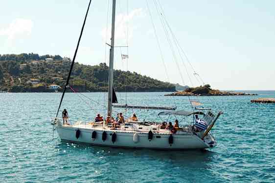 En seilbåt med flere personer om bord seiler nær en kystlinje med grønne åser og noen hus. (AI-generert tekst)