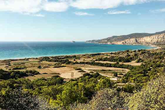 Et landskapsbilde med grønne marker, busker og fjell som strekker seg mot en strand med turkis hav under en klar himmel. (AI-generert tekst)