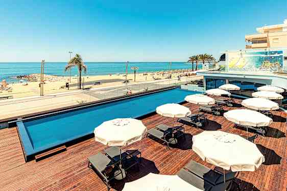 Et utendørs svømmebasseng med solsenger og parasoller, og en sandstrand med havutsikt. (AI-generert tekst)