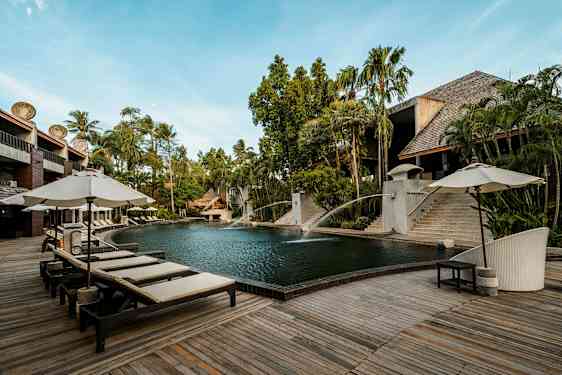 Et utendørs svømmebasseng omgitt av tredekke og solsenger med parasoller. Det er frodig vegetasjon og bygninger av tre og stein i bakgrunnen. (AI-generert tekst)