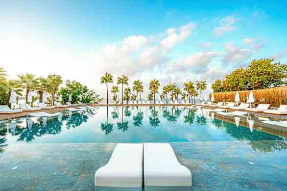 Et utendørsbasseng med solsenger og parasoller, omgitt av palmer, og med speilinger i vannet. (AI-generert tekst)
