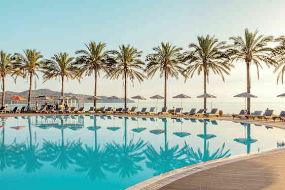 Et basseng med solsenger og parasoller, omgitt av palmer. Havet er synlig i bakgrunnen. (AI-generert tekst)