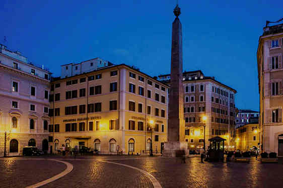Et torg med bygninger rundt og en obelisk i midten, opplyst om natten. (AI-generert tekst)