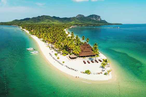 En tropisk øy med palmer og hvite sandstrender omgitt av turkist hav. En bygning med mørkt tak er synlig på stranden. (AI-generert tekst)