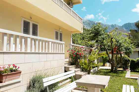 Bilde av en husfasade med balkong og hage i forgrunnen, inkludert benker og blomster. (AI-generert tekst)