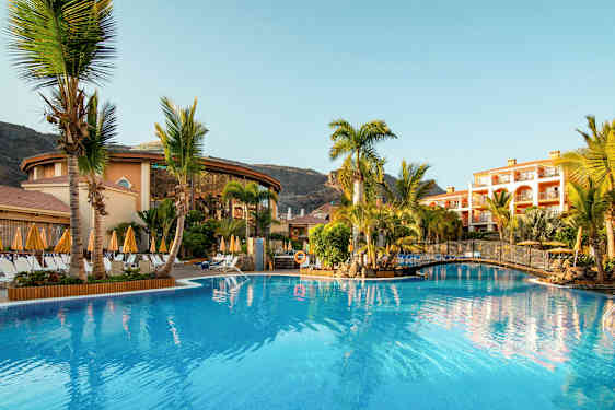 Bilde av et hotellområde med et basseng, omgitt av palmer og bygninger. (AI-generert tekst)