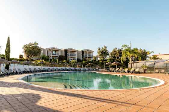 Utendørs svømmebasseng med solsenger og parasoller, omgitt av grøntområder og bygninger. (AI-generert tekst)