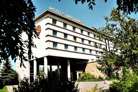 Bygning med flere etasjer, merket med teksten 'Hotel Sympozjum', omgitt av trær og vegetasjon. (AI-generert tekst)