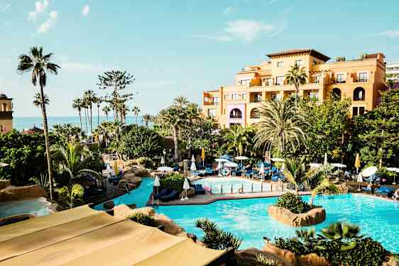 Hotellområde med bassenger og grøntområder foran en bygning, hav i bakgrunnen. (AI-generert tekst)