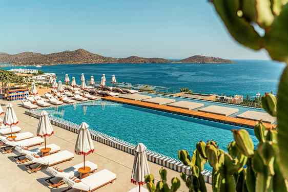 Et basseng med solsenger og parasoller ved en kystlinje med utsikt over en blå sjø og fjell. (AI-generert tekst)