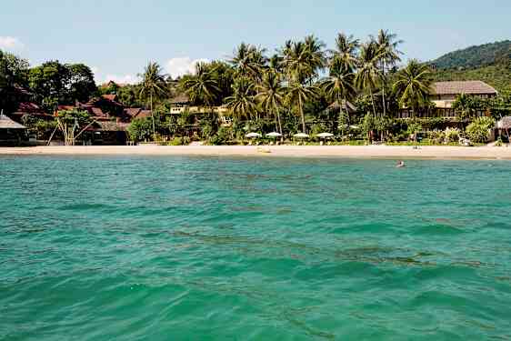 Utsikt over en sandstrand omgitt av tropiske trær og bygninger, sett fra vannet. (AI-generert tekst)