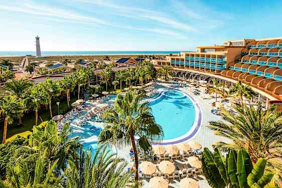 Et hotellanlegg med et stort basseng, solsenger og parasoller omgitt av palmer, med sjøen og et fyrtårn i bakgrunnen. (AI-generert tekst)