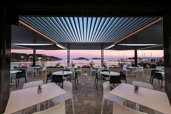 En utendørs terrasse med bord og stoler med utsikt mot havet og seilbåter. (AI-generert tekst)