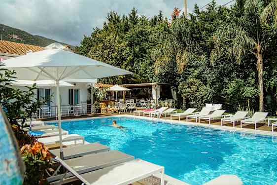 Et utendørs basseng omgitt av hvite solsenger og parasoller nær en bygning og grøntområder. (AI-generert tekst)