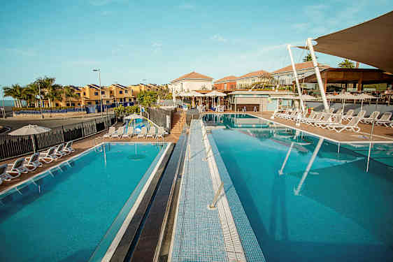 Et bilde av et solrikt feriested med to utendørsbassenger omgitt av solsenger, med bygninger i bakgrunnen. (AI-generert tekst)