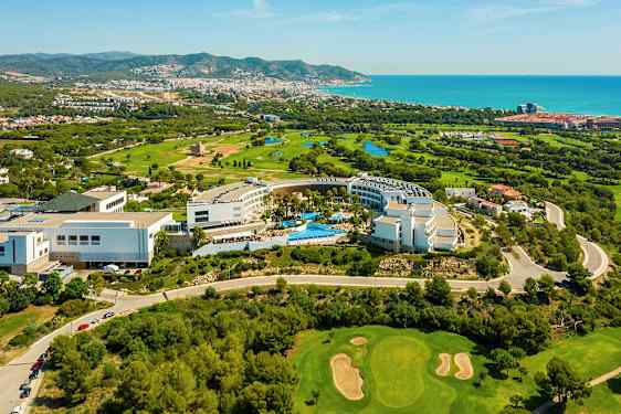 Utsikt ovenfra over et hotellområde omgitt av grøntområder og en golfbane. Havet og en by er i bakgrunnen. (AI-generert tekst)