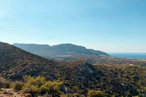 En utsikt over et fjelllandskap med variert vegetasjon og havet i bakgrunnen under en klar blå himmel. (AI-generert tekst)